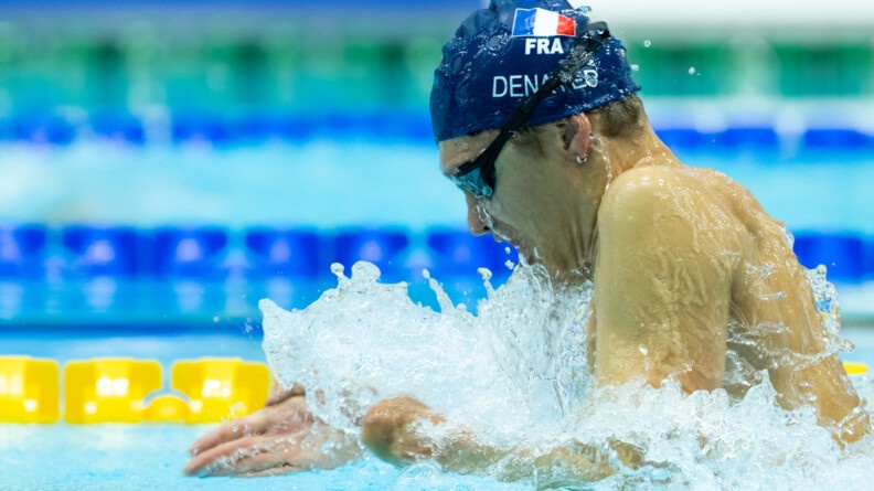 De profil, sortant de l'eau faisant une vague autour de lui, une garçon portant des lunettes de natation noires et un bonnet bleu marine siglé d'u drapeau français et du nom Denayer, s'apprête à replonger dans l'eau