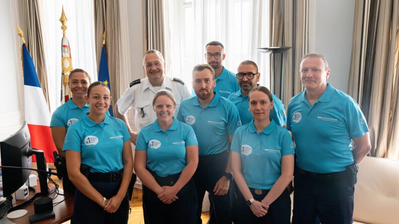 Dans un bureau, le directeur général de la gendarmerie pose au milieu de huit gendarmes vêtus d'un polo bleu turquoise. A leur gauche, on aperçoit un bureau et un grand drapeau français