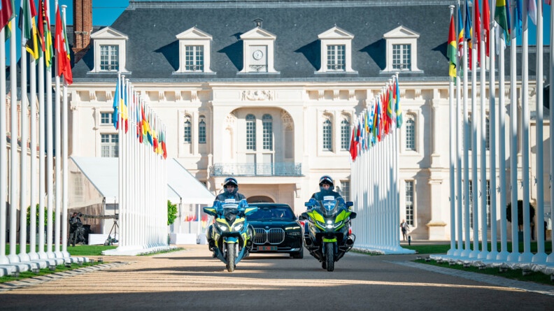 Dans une marge allée, devant un château, une berline noire avance, précédée de deux gendarmes motocyclistes. De nombreux drapeaux de différents pays, non hissés, jalonnent les bords de l'allée.