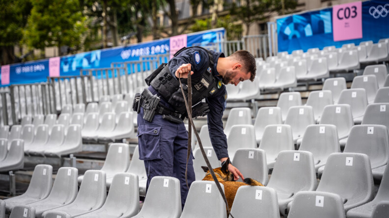 Au milieu de fauteils gris, un gendarme en combinaison bleue marine et gilet par-balles noir montre à son chien un fauteuil