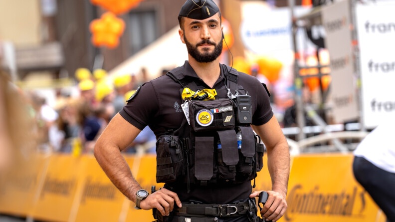 Un gendarme, revêtant un uniforme noir, se tient debout sur la route, mains sur les hanches, face à l'objectif. Derrière lui on aperçoit une grande bannière jaune, sur laquelle est inscrit "CONTINENTAL"