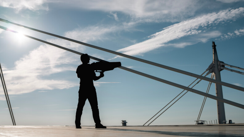 Sur le toit du Stade de France, un homme se tient debout, de dos, face à un grand ciel bleu parsemé de nuages blancs, un fusil anti-drone dans les mains.