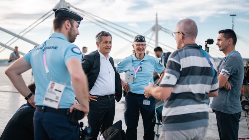 Sur le toit du Stade de France, deux gendarmes et deux hommes en civil discutent avec Gérald Darmanin, ministre de l'intérieur et des outre mer. Ce dernier se tient debout, face à l'objectif, les mains posées sur les hanches