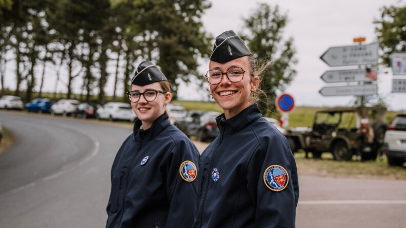 Deux jeunes filles portant la tenue des cadets de la gendarmerie du Calvados, souriantes. En arrière-plan, floues, des voitures stationnées le long de la route