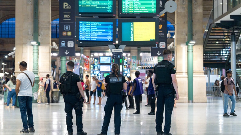 Trois gendarmes mobiles de dos, dans un hall de gare, scrutent la foule en transit que l'on voit en arrière-plan. Au-dessus d'eux quatre écrans d'affichage SNCF.