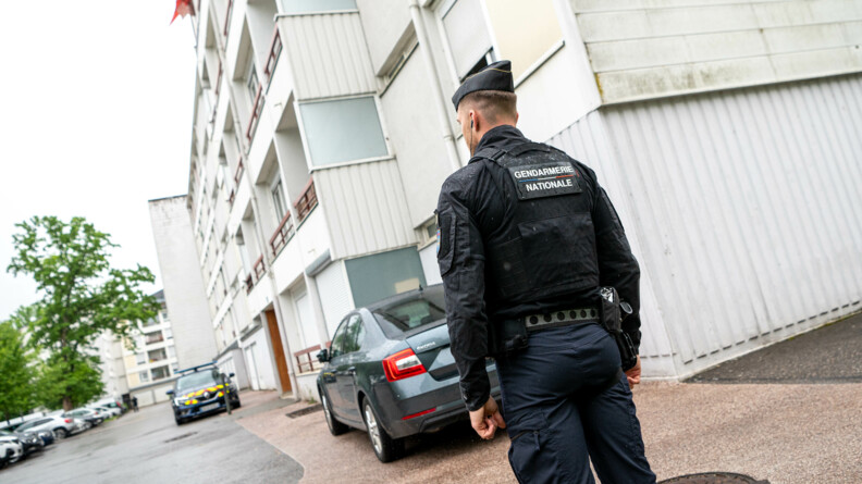 Un gendarme en tenue se tient debout, de dos, devant l'immeuble d'une cité. Son blouson porte dans le dos la mention "Gendarmerie nationale".