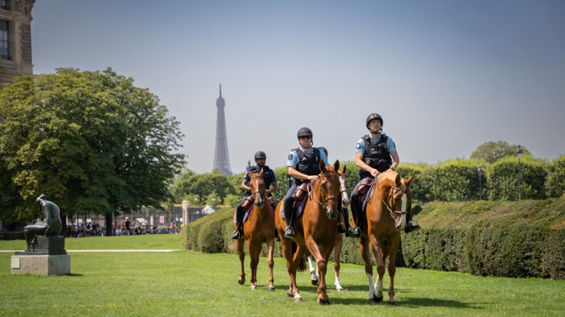 Quatre cavaliers, marchant deux par deux, sur l'herbe, devant des haies et arbres, ainsi qu'une statue à gauche. En fond, la Tour Eiffel