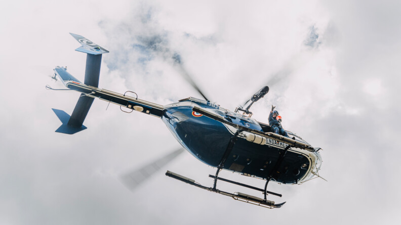 Vue en contre plongée d'un  hélicoptère gendarmerie dans le ciel nuageux, un gendarme à la porte latérale lançant un lest
