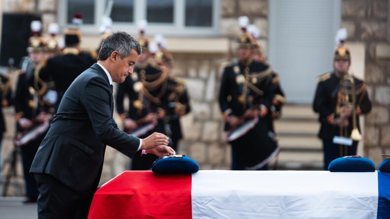 Un homme de profil en costume foncé place un objet sur le coussin bleu posé sur un cercueil recouvert d'un drapeau tricolore. En arrière-plan flou, des musiciens de la Garde républicaine