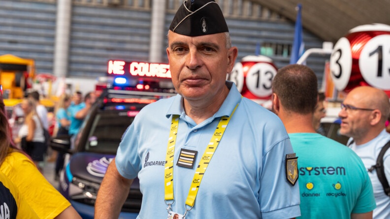 Une gendarme se tient debout face à l'objectif ; il porte un tour de cou jaune sur lequel est écrit en lettres noies Tour de France. derrière lui se trouvent des personnes qui discutent, ainsi qu'un véhicule de la gendarmerie portant une bannière lumineuse sur le toit, diffusant des messages de prévention ("Ne courez pas")