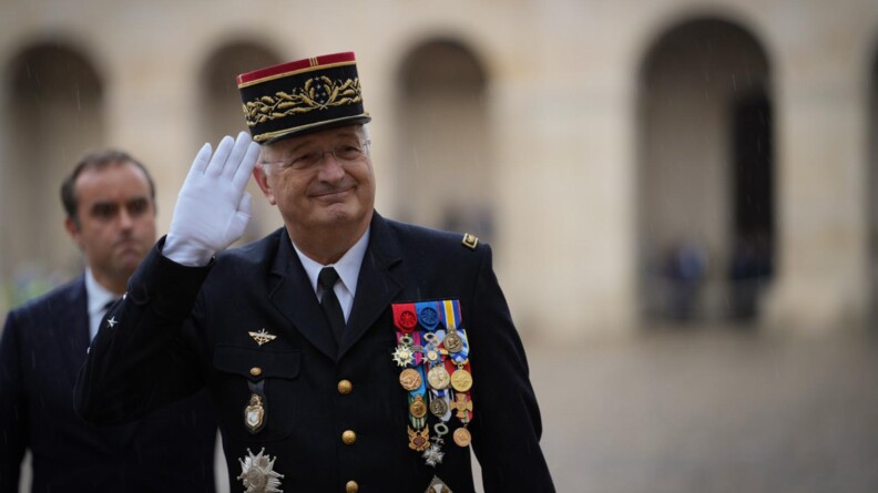 Au premier plan, le directeur général de la gendarmerie nationale, en buste, esquisse un sourire en effectuant un salut de sa main droite gantée. Au second plan, à gauche, flou, le ministre des Armées le suit.