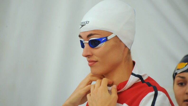 Une femme, vue en buste, de profil gauche, porte ses main à son cou. Elle porte une veste de sport blanche et rouge, des lunettes de piscine bleu et blanche et un bonnet de bain blanc