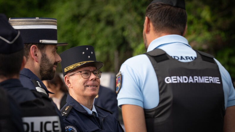 Le Major Général de la Gendarmerie Nationale échange, souriant,  avec un gendarme, que l'on voit de dos. Ce dernier porte un polo bleu ciel et un gilet pare balles noir revêtant l'inscription "Gendarmerie" dans le dos. Un colonel de la gendarmerie se tient debout à leurs côtés.