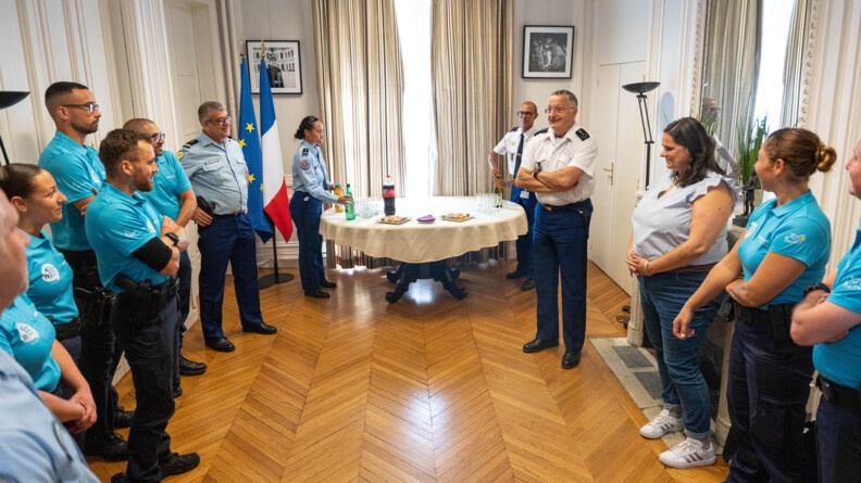 Dans un bureau, le directeur général de la gendarmerie pose au milieu de gendarmes vêtus d'un polo bleu turquoise et d'une femme brune portant une tenue civile. Au fond de la pièce une table est dressée sur laquelle sont posés des bouteilles et quelques petits gâteaux. On aperçoit dans l'un des angles de la pièce un grand drapeau français ainsi qu'un grand drapeau européen.