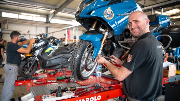 Dans un atelier de réparation automobile, un mécanicien réparant une moto bleue de la gendarmerie, se tourne, souriant, vers l'objectif. L'homme est vêtu d'un t-shirt et d'un pantalon noirs.