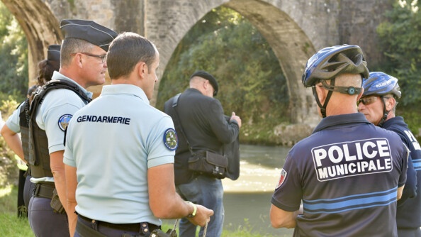 TRois gendarmes, deux policiers municipaux et un homme en jean veste noire au bord d'un cours d'eau surmonté d'un pont