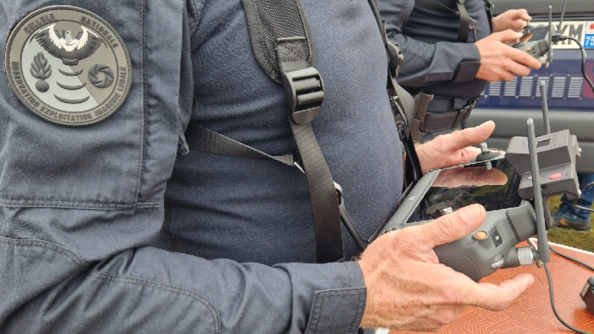 Deux gendarmes, debout, de biais, dont on voit le tronc et les mains, tiennent dans leurs mains une manette, comprenant un écran et des commandes