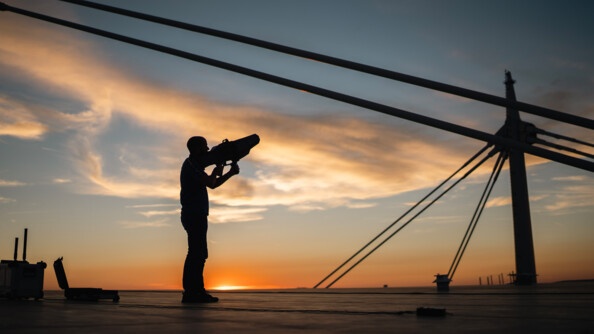 Sur le toit du Stade de France, un homme se tient debout, de biais, face au ciel où le soleil se couche, un fusil anti-drone dans les mains.