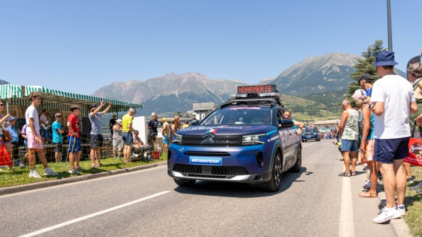 Un véhicule bleu marine, portant l'inscription GENDARMERIE sur la portière, en grosses lettres blanches, roule sur la route. Une bannière lumineuse, posée sur le toit, délivre des messgaes de prévention. Des spectateurs se tiennent debout, sur le bord de la route. L'un d'eux agite les mains au passage de la voiture. On aperçoit la montagne en arrière plan. Le ciel est bleu et ensoleillé.