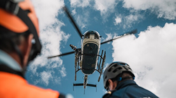 Vue en contre-plongée d'un hélicopète gendarmerie dans le ciel nuageux, présence floue en bas de l'image de deux gendarmes dont un porte une chasuble orange