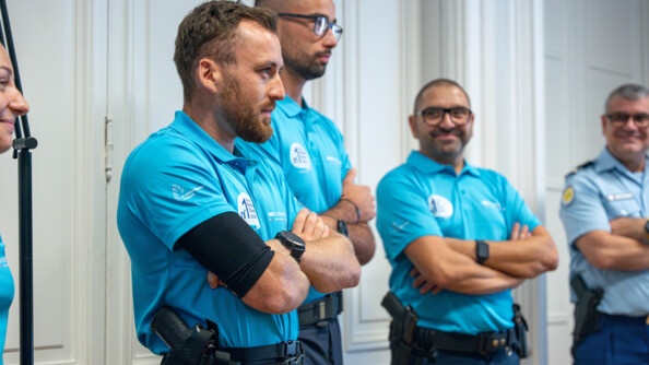 Trois gendarmes vêtus d'u  pantalon bleu marine et d'un polo bleu turquoise se tiennent debout, les bras croisés contre leur ventre, regardant devant eux.