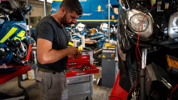 Dans un atelier de réparation automobile, un mécanicien se tient debout, à côté d'une moto noire. L'homme est vêtu d'un t-shirt et d'un pantalon gris, et il tient dans sa main un outil que l'on devine, mais que l'on ne voit pas.