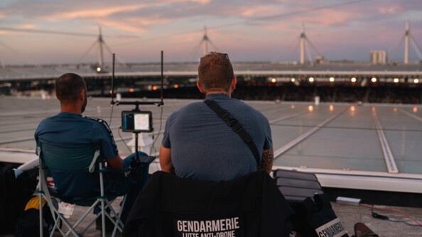 Sur le toit du Stade de France, on voit deux hommes de dos, assis sur des chaises, face au ciel. Sur le dossier de la chaise de l'un des deux hommes, une veste noire est posée qu porte l'inscription Gendarmerie lutte anti-drone