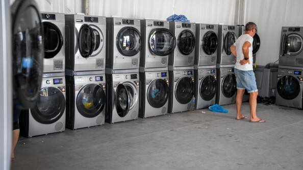 Photo d'une buanderie équipée de nombreux sèches-linges et machines à laver, superposés les uns sur les autres. Un homme, vêtu d'un t-shirt blanc et d'un short en jean, se tient debout, de dos, les mains sur les hanches, face à l'une des machines