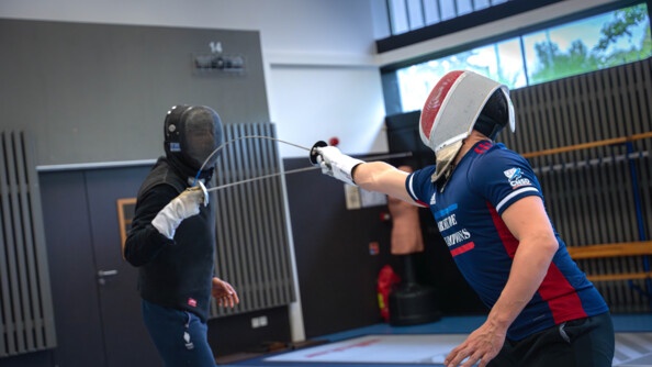 A fencer dressed in the tracksuit of the army of champions touches his fencing master in front of him, dressed in black, who does not parry