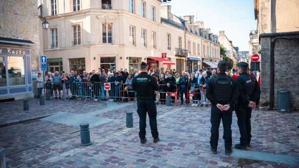 Trois gendarmes sécurisent un barrièrage derrière lequel les spectateurs observent la cérémonie. Le tout dans les rues pavées de Bayeux.