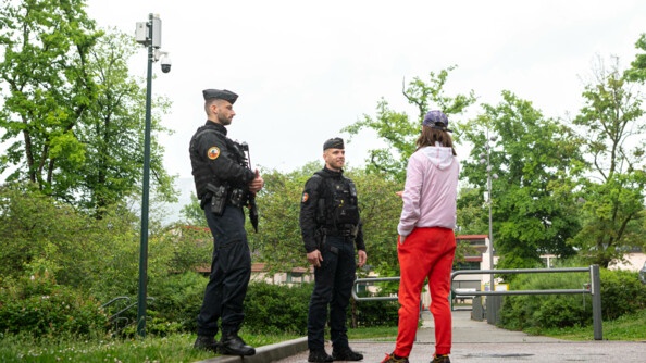 Deux gendarmes discutent avec un adolescent portant une casquette violette, un blouson rose, un jogging rouge et des baskets. lls se tiennent debout, dans l'espace extérieur et verdoyant d'une résidence