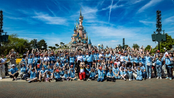 150 jeunes vêtus de bleu ciel, posent en levant les mains en l'air. En arriève-plan, le château de la belle au bois dormant de Disneyland Paris