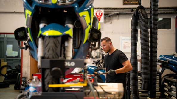 Dans un atelier de réparation automobile, un mécanicien répare une moto bleue de la gendarmerie. L'homme se tient debout, face à l'objectif. Il est postionné derrière la moto, et est vêtu d'un t-shirt noir et de gants noirs..