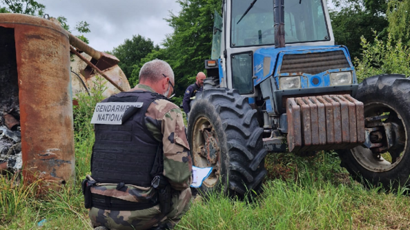 deux gendarmes inspectent un tracteur abandonné en pleine nature. l'un d'eux est accroupi, un genou à terre, devant l'engin, et prend des notes sur un support