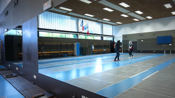 In the reflection of the wall mirror, two fencers confront each other in a weapons room