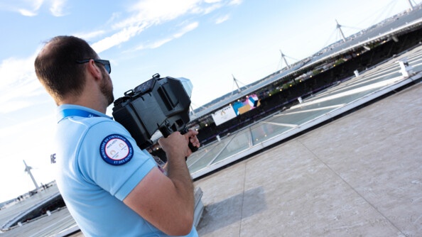 Sur le toit du Stade de France, un homme se tient debout, de dos, face à une grand ciel bleu parsemé de nuages blancs, un fusil anti-drone dans les mains.