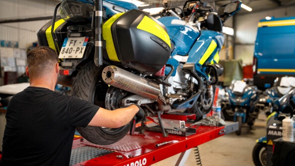 Un homme vêtu d'un t-shirt noir, et de gants noirs, se tient debout de dos, en premier plan. Il répare une moto de la gendarmerie positionnée sur un élévateur. L'homme se trouve dans un atelier de réparation automobile.