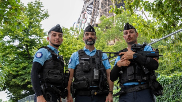 Trois gendarmes se tiennent debout, côte à côte, devant la Tour Eiffel. L'un d'eux, situé à droite, tient dans ses mains un fusil.