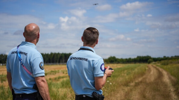 Deux gendarmes en pantalon bleu foncé, polo bleu clair manches courtes, vus de dos, plan en buste. Ils sont dans un champ ; en fond un bois. Celui de droite a une télécommande dans les mains. Ils regardent devant eux, légèrement vers le ciel, où vole un drone.