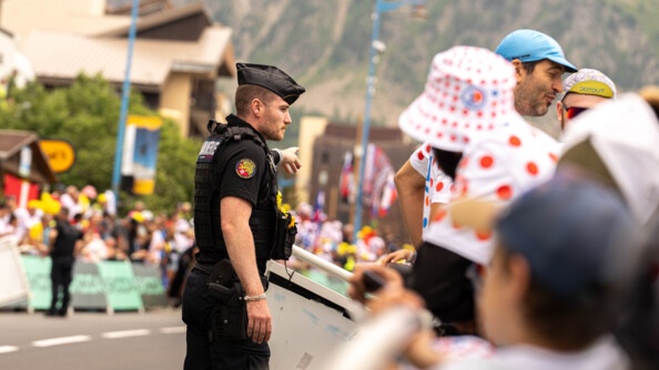 Un gendarme, revêtant un uniforme noir, se tient debout sur la route face à une foule. Il désigne un point avec l'index de sa main gauche. On aperçoit la montagne en arrière plan.