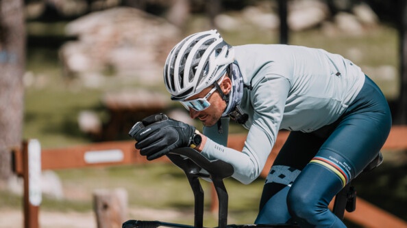 Un cycliste avance sur son vélo, le corps penché en avant. Vêtu d'une tenue bleue, il porte un casque, des lunettes de soleil blanches et des gants noirs.