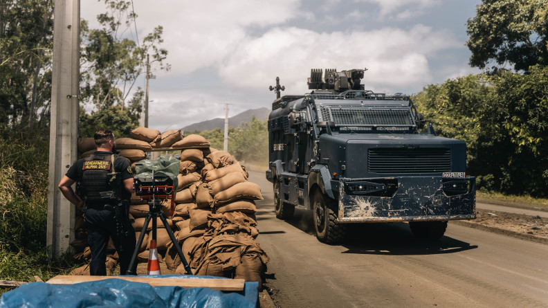 Agrandir l'image Un gendarme mobile vu de dos, derrière un mur de sacs de sable. Devant lui, sur la route, un Centaure.